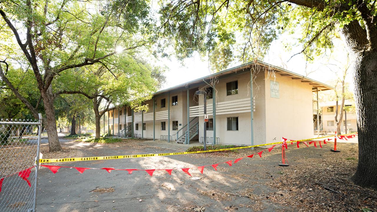 Image of 1970s era apartment building surrounded by large trees. In the forefront there's a line of yellow caution tape.